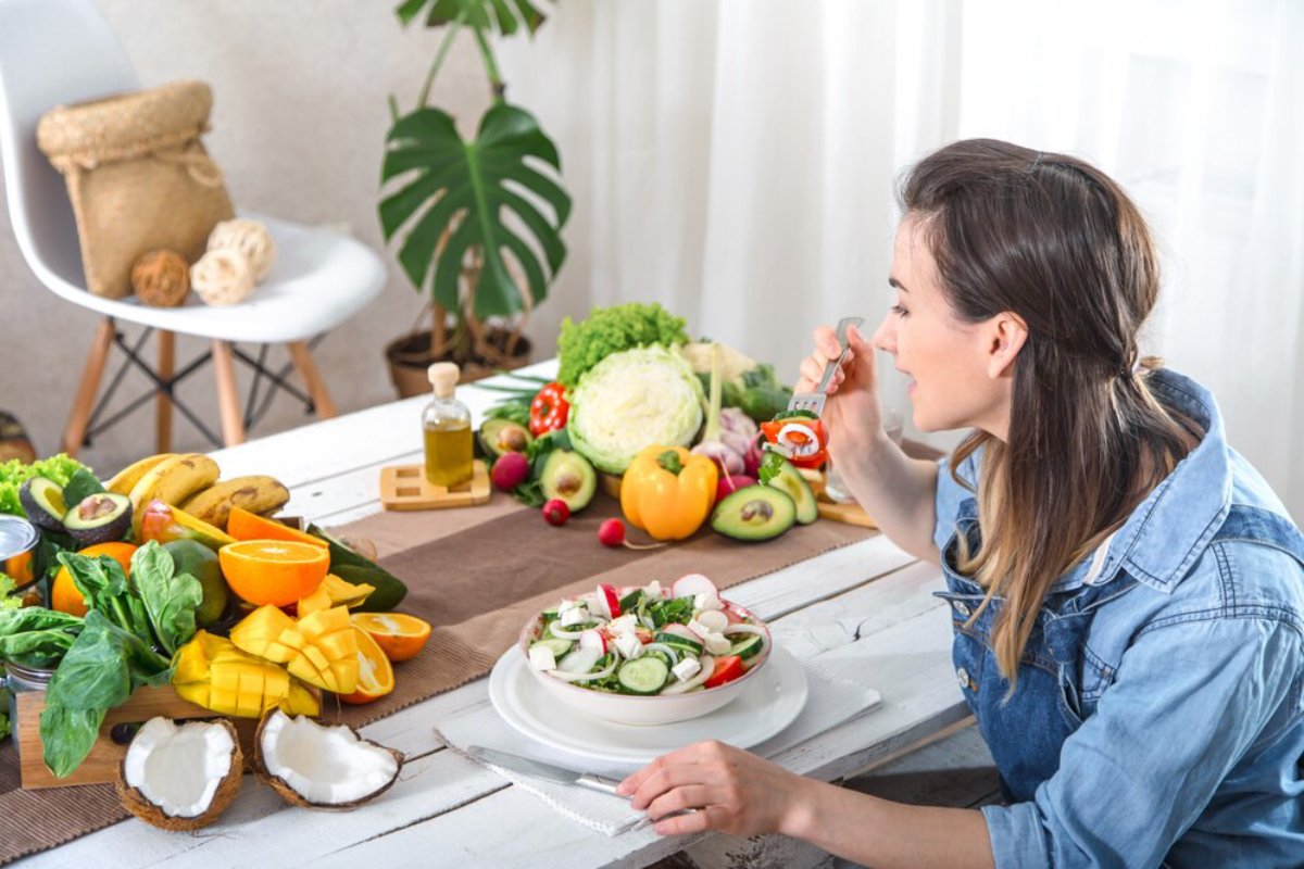 Woman eating a salad at a table full of fresh fruits and vegetables in a bright kitchen.