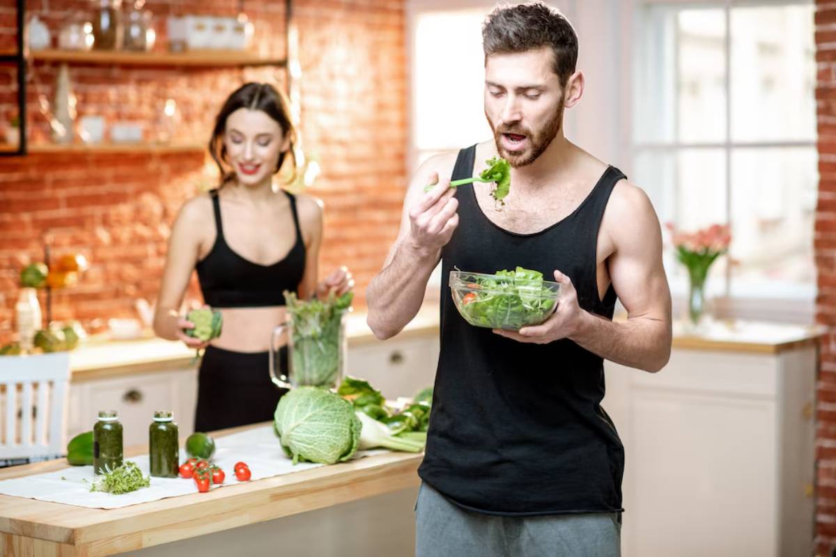 A man eats a salad while a woman prepares vegetables in a stylish kitchen.