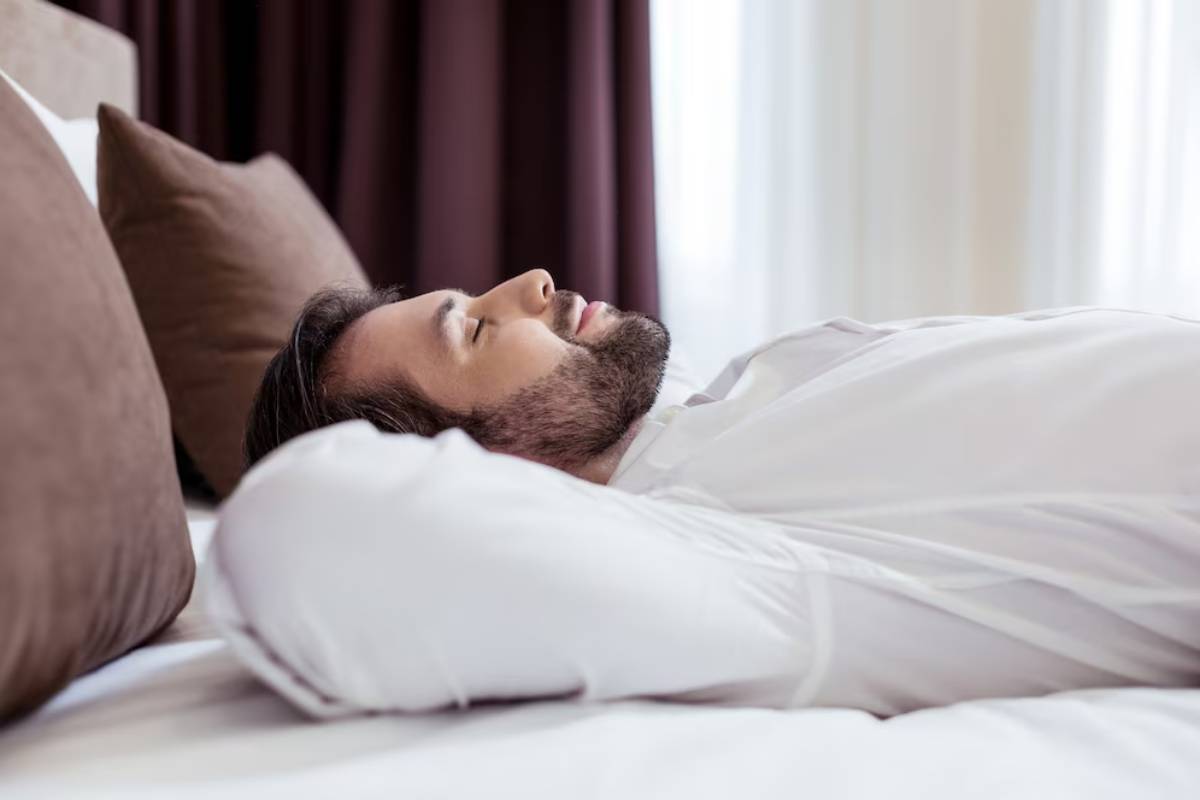 Man with beard sleeping peacefully in a white bed in a well-lit room.