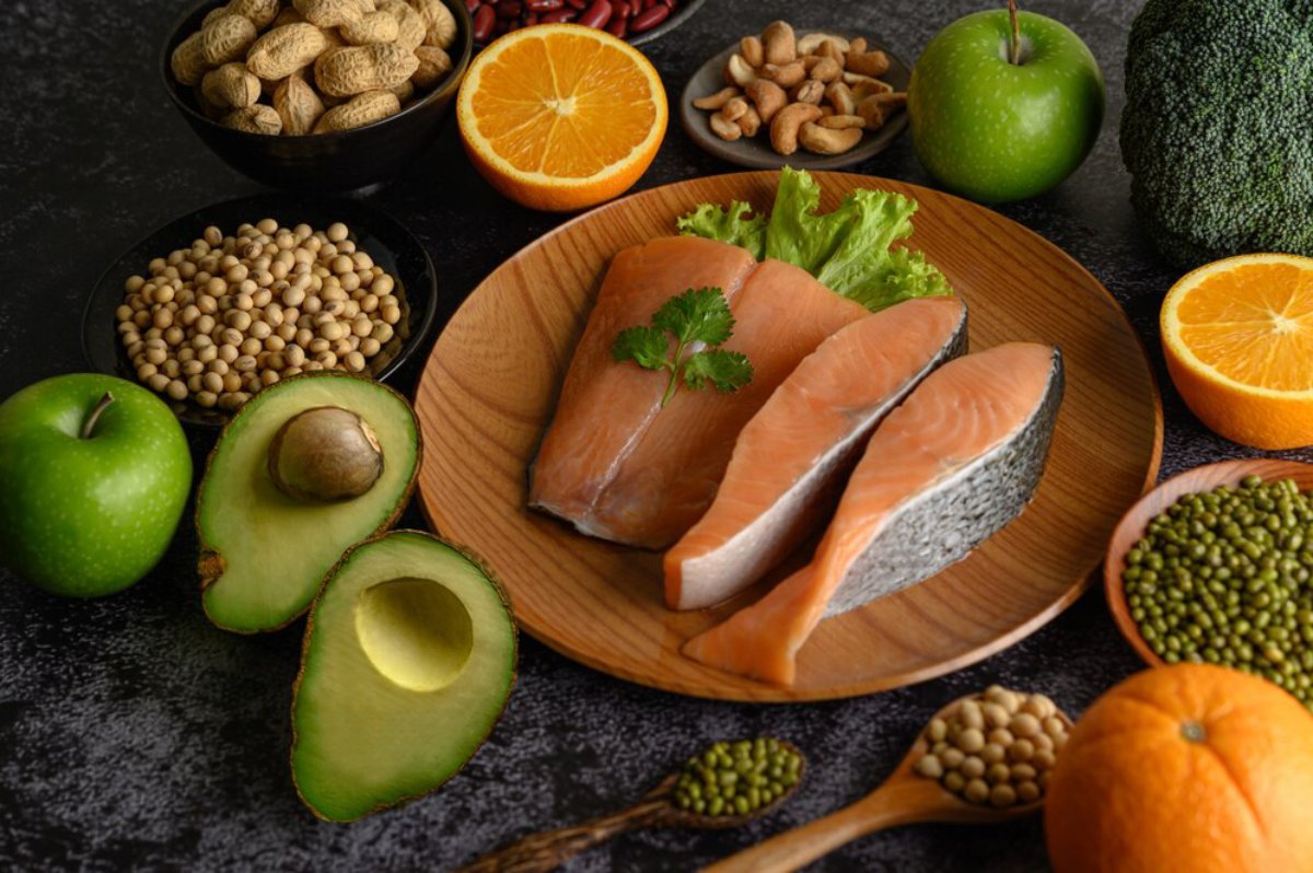 A variety of healthy foods including salmon, avocados, nuts, and fresh fruits displayed on a dark surface.