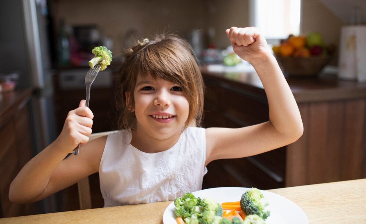 Young girl at kitchen table happily holding broccoli on a fork, flexing her arm muscles.