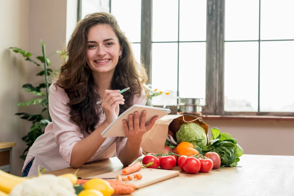 Young woman smiling and reading a recipe on a tablet amidst fresh vegetables on a kitchen counter.