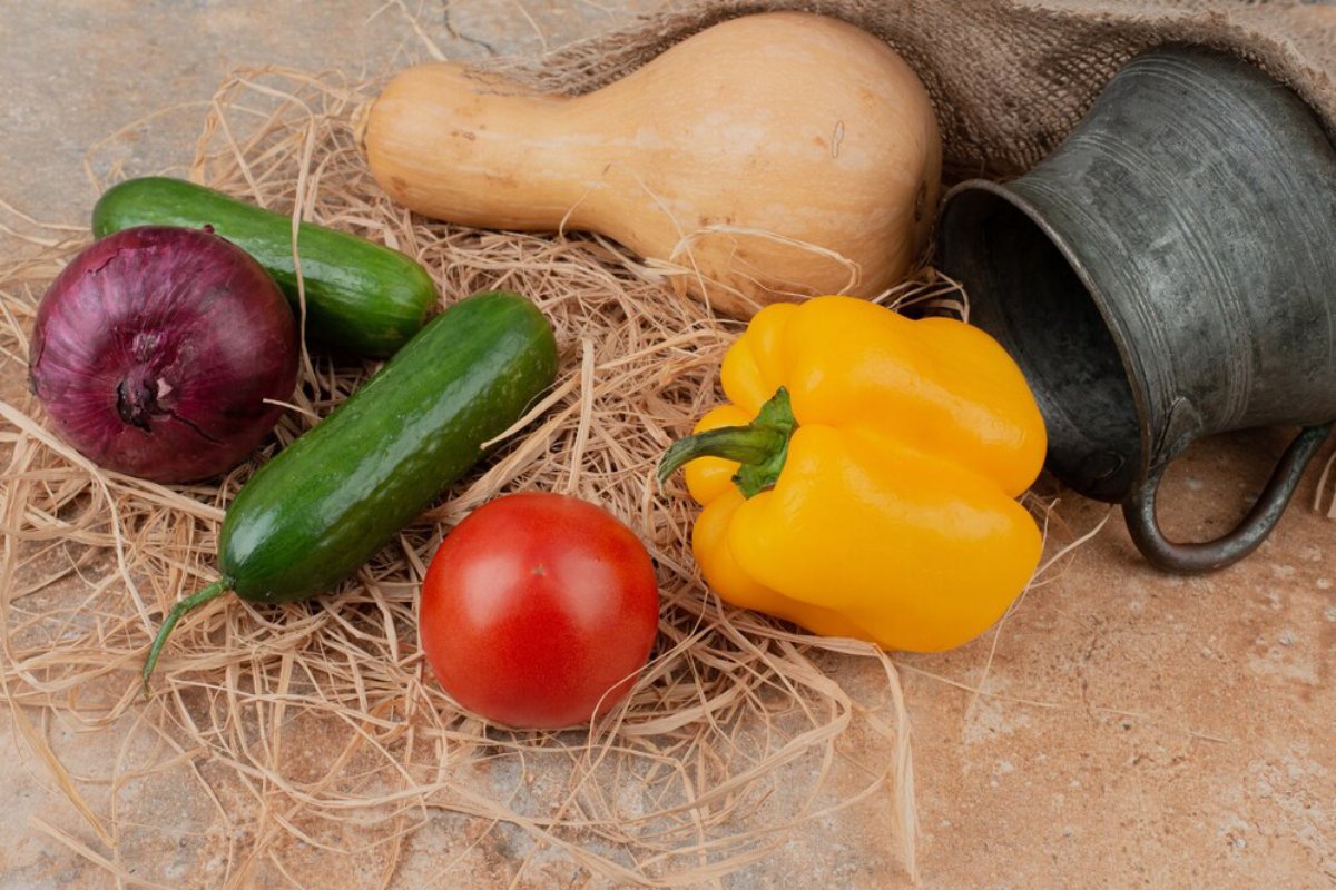 Various fresh vegetables including a butternut squash, cucumbers, a tomato, a red onion, and a yellow bell pepper on straw.
