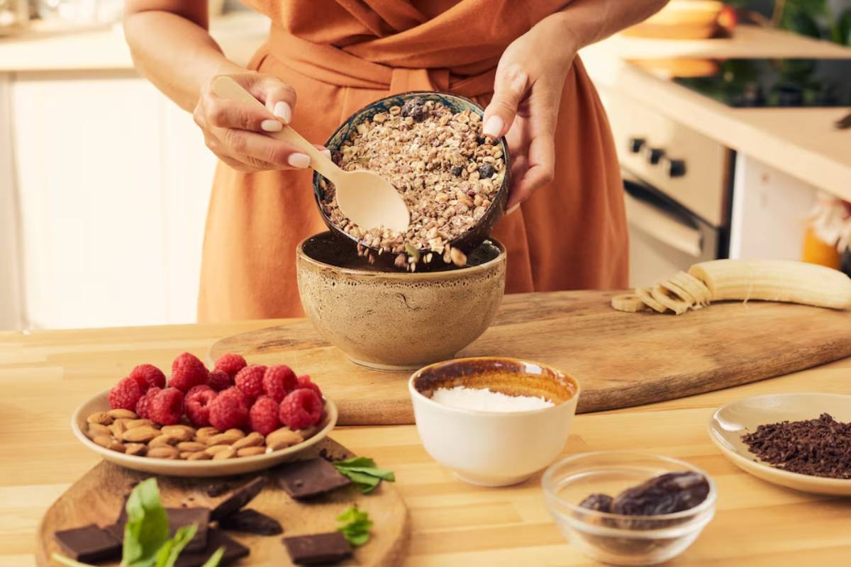 Person preparing a healthy meal by adding granola to a bowl in a well-equipped kitchen, with ingredients like raspberries and almonds on the side.
