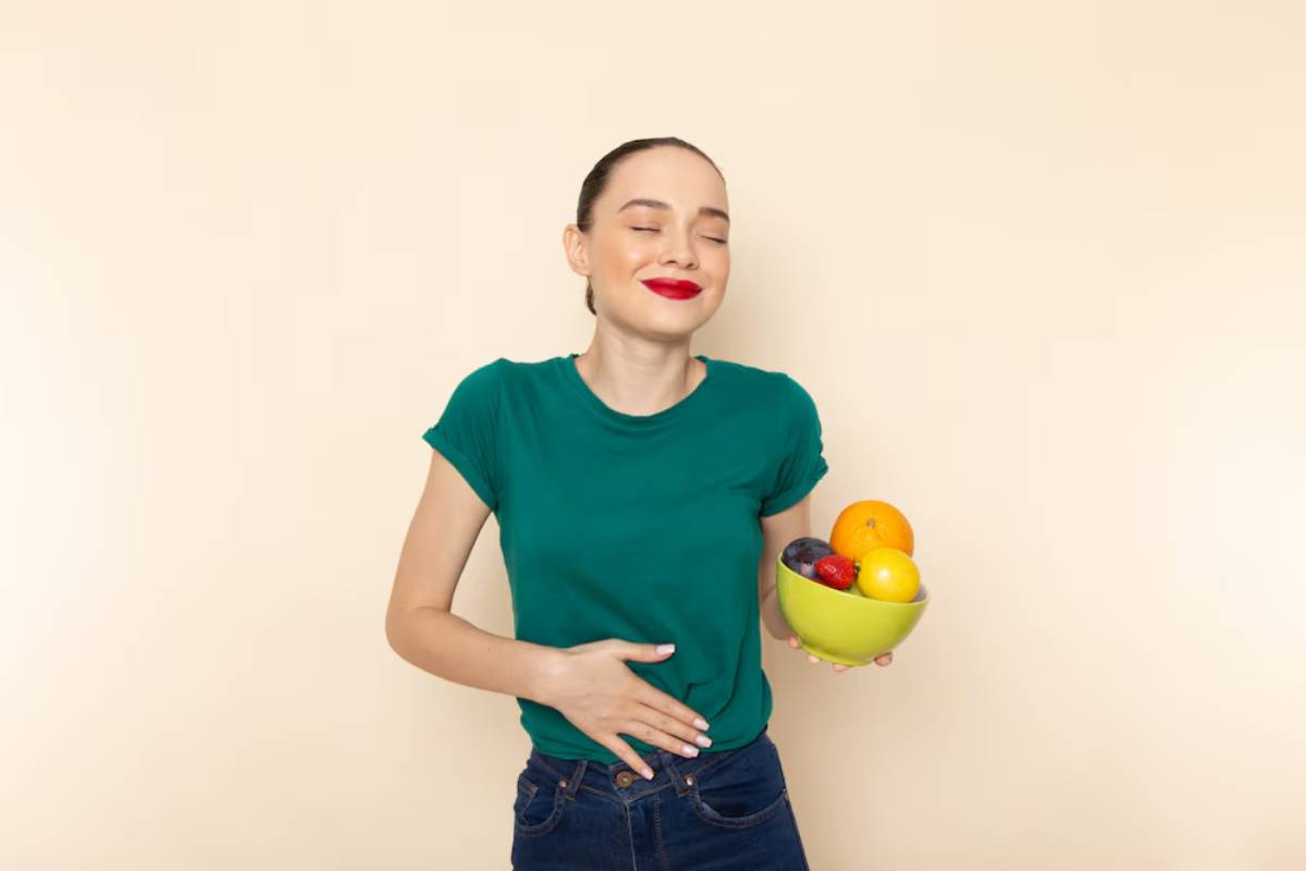 Woman in green t-shirt smiling with closed eyes, holding a bowl of fruit against a light beige background.
