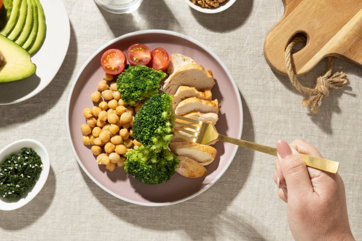 A hand holding a fork over a healthy meal with chicken, broccoli, chickpeas, and tomatoes on a plate.