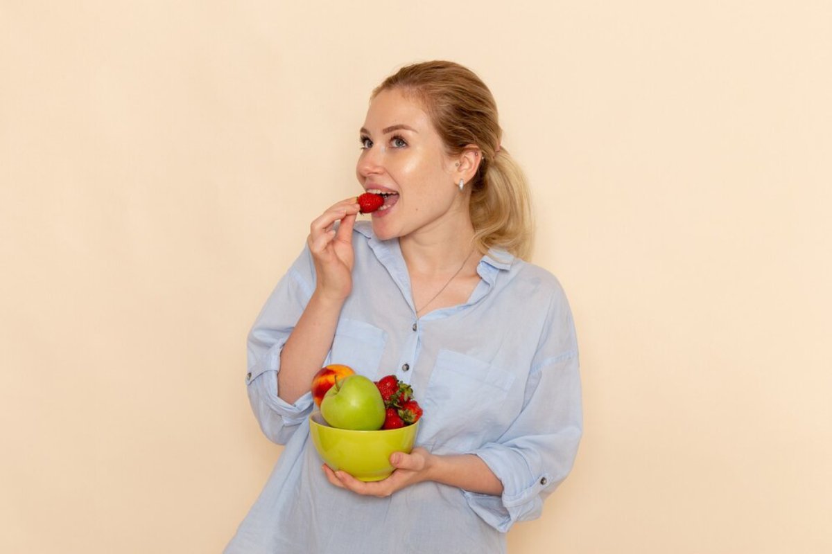 Woman in a blue shirt eating a strawberry and holding a bowl of mixed fruit against a light beige background.