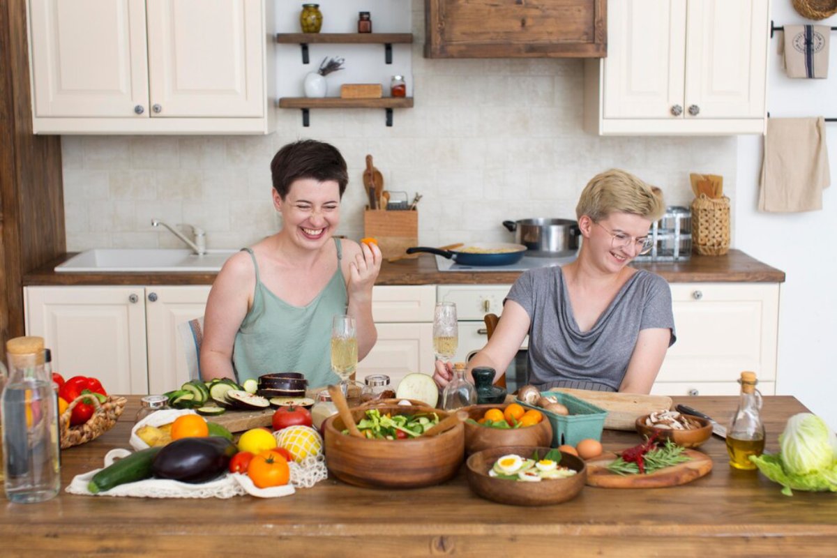 Two women laughing and preparing food together in a bright kitchen filled with fresh vegetables and wooden bowls.