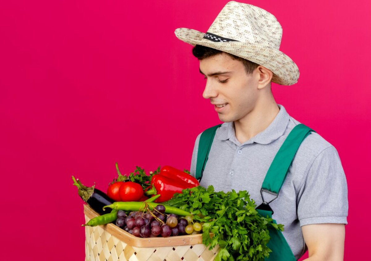 A young man in a straw hat looks at a basket full of fresh vegetables against a pink background.