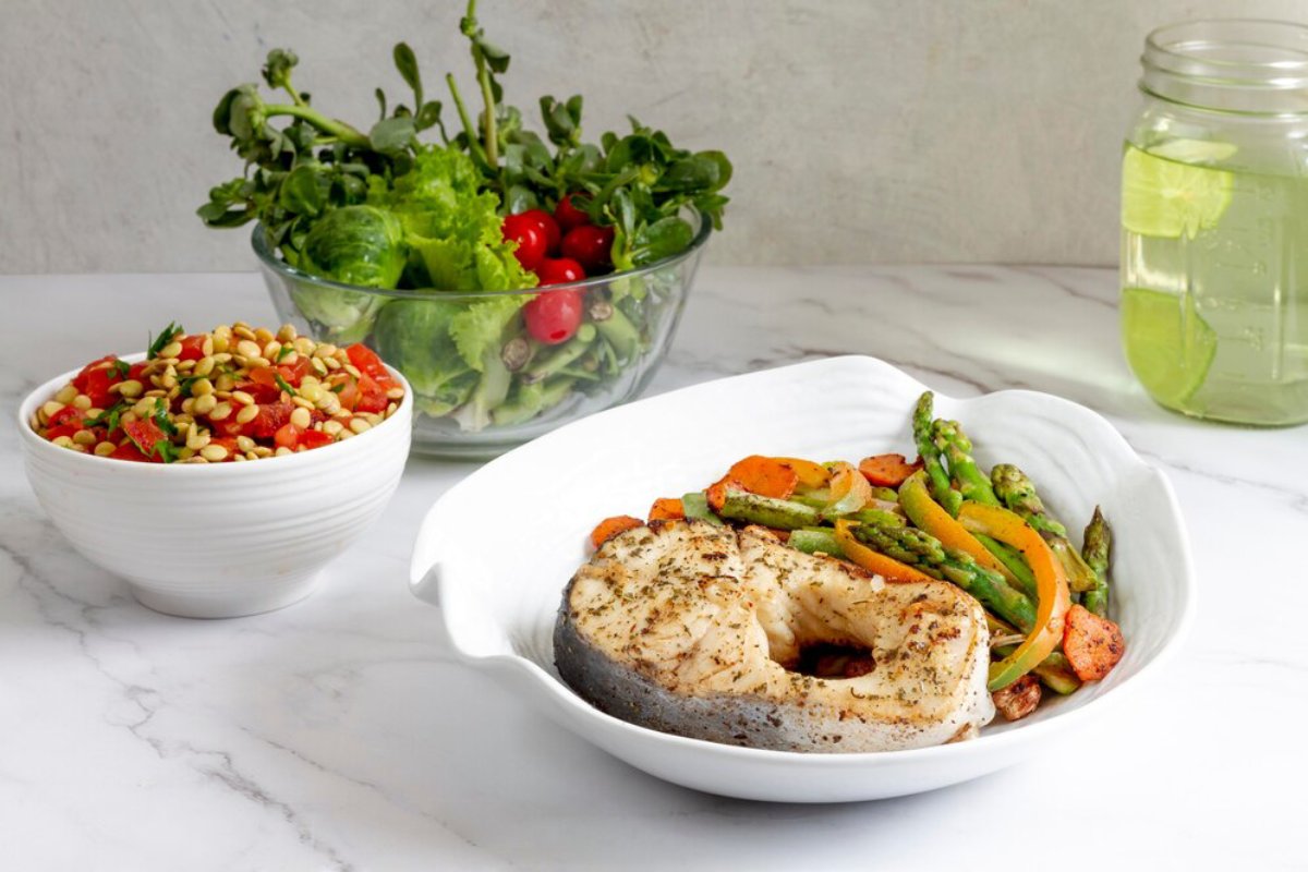 Grilled fish with vegetables on a white plate, accompanied by salad and a bean dish, with a drink in the background.