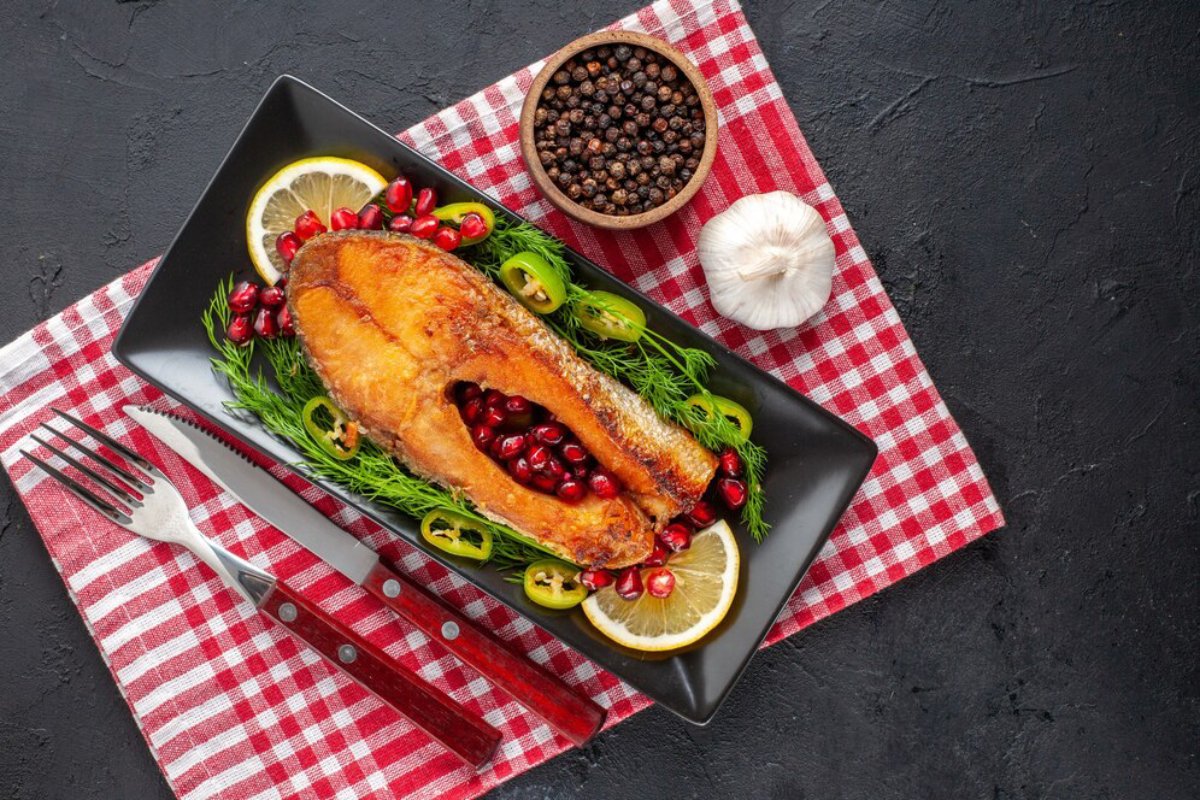 Roasted fish garnished with lemon, pomegranate seeds, and herbs on a black plate with red checkered napkin beside.