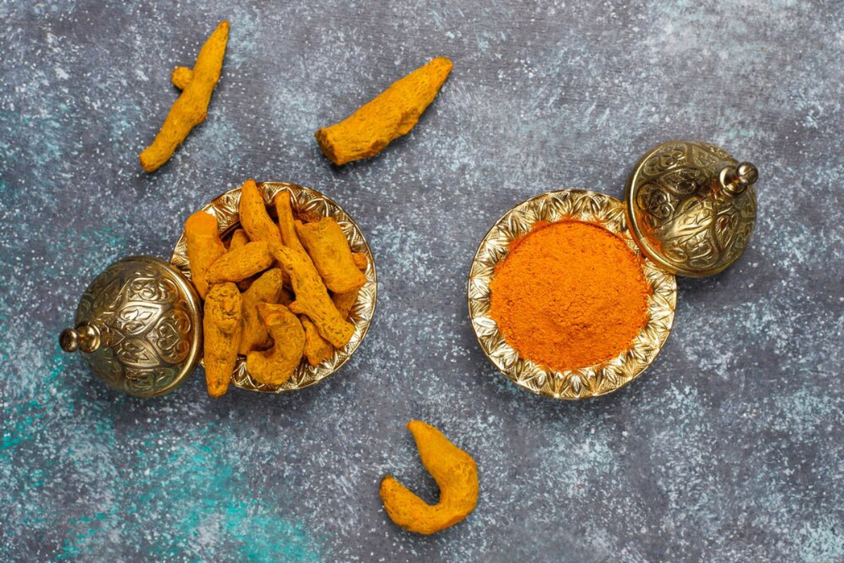 Turmeric roots and powder in decorative metal dishes on a textured blue surface.