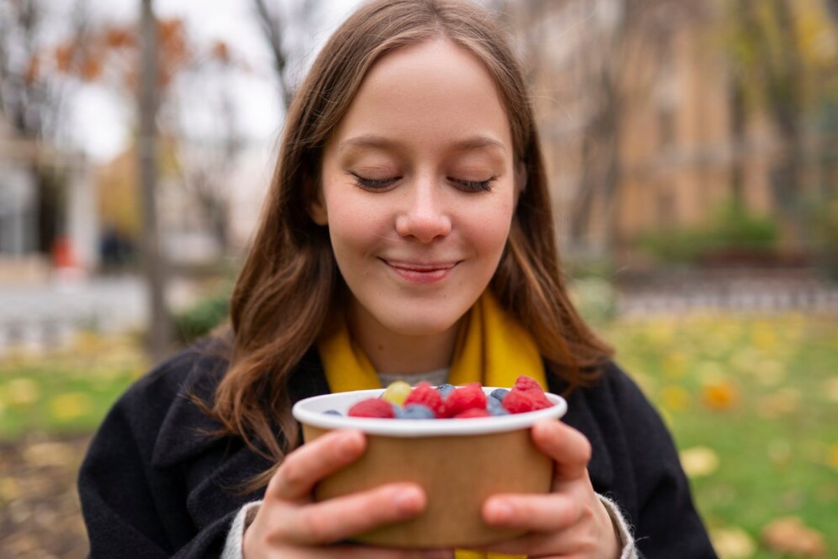 Young woman smiling, holding a bowl of fresh mixed berries in a park.