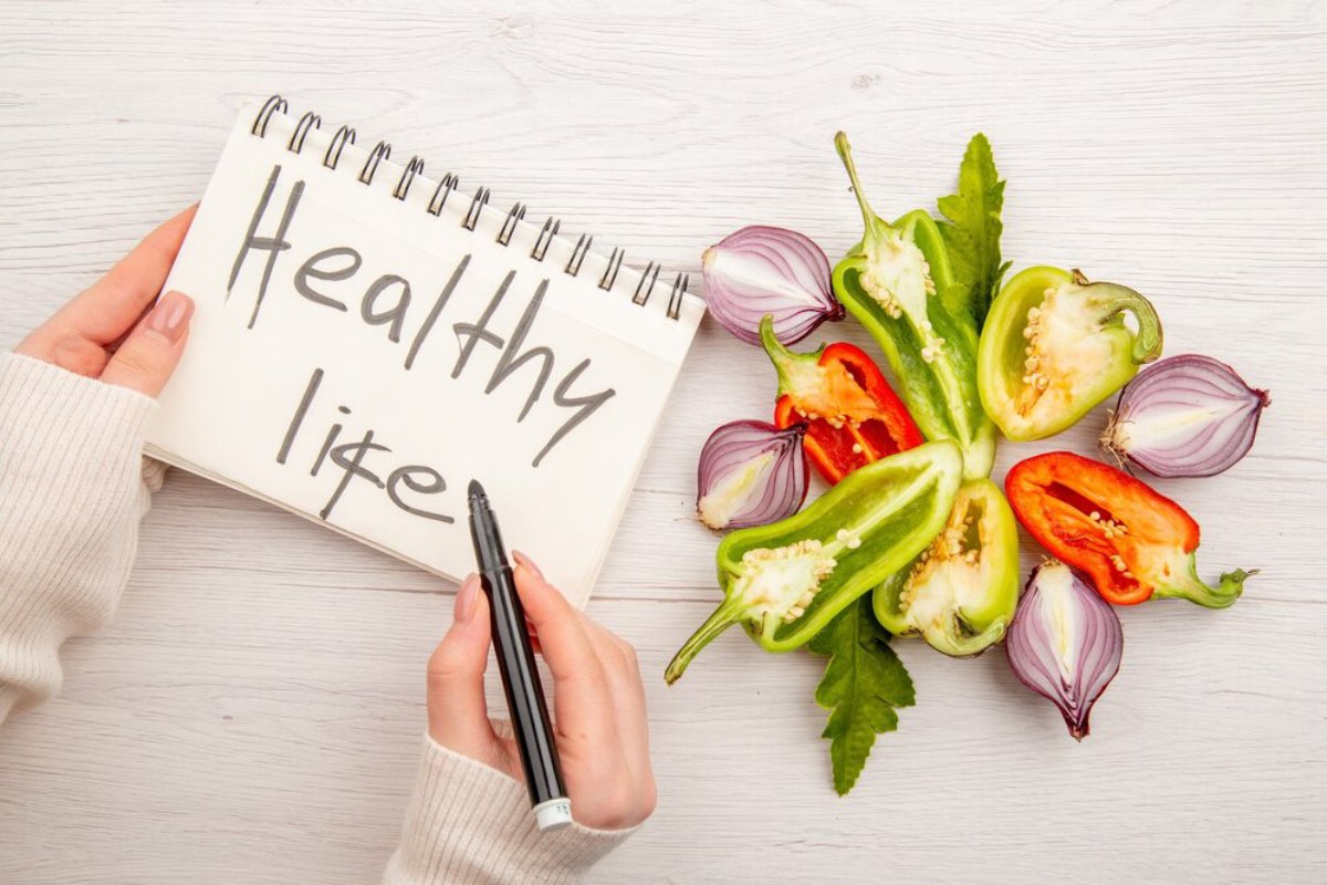 Person writing Healthy Life in notebook beside fresh bell peppers and onions on a wooden table.