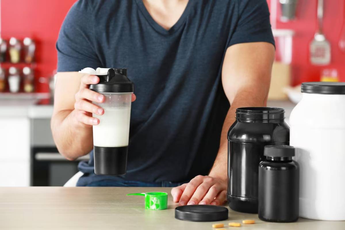 Man holding a protein shake with supplement containers and a scoop on a kitchen counter.