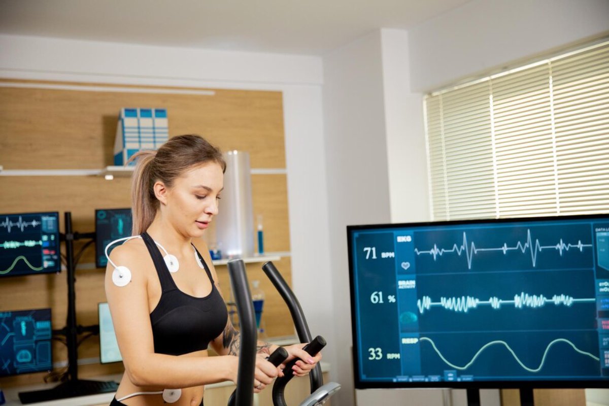 A woman uses an elliptical machine in a lab, monitored by multiple screens displaying her biometric data.