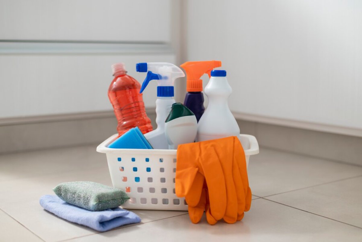 A basket of various cleaning supplies including bottles, gloves, and cloths on a tiled floor.