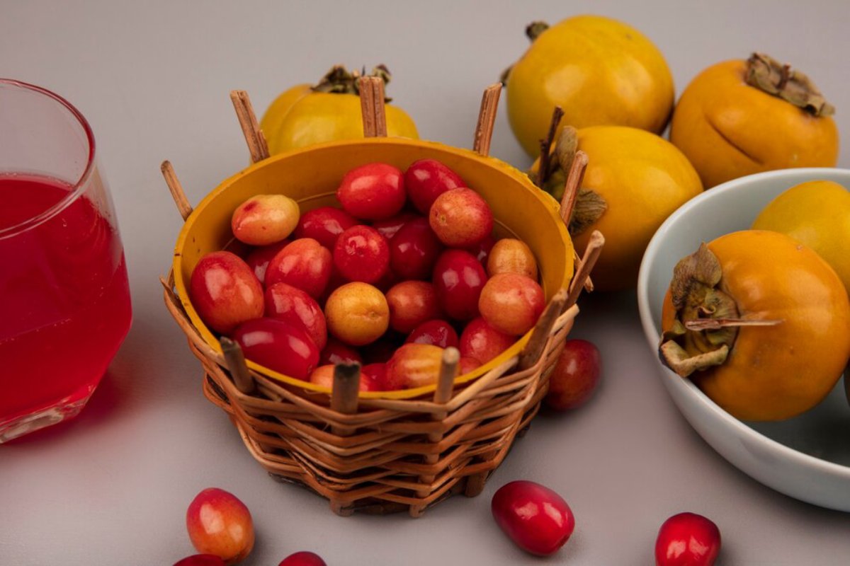 A wicker basket filled with colorful cranberries, flanked by persimmons and a glass of red juice on a gray background.