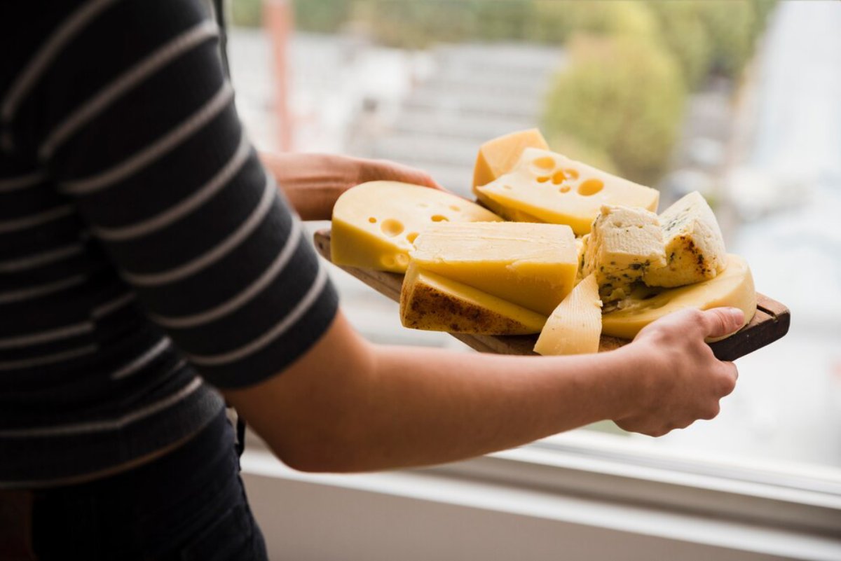 Person holding a wooden board with assorted cheeses, near a window, with a blurred outdoor background.