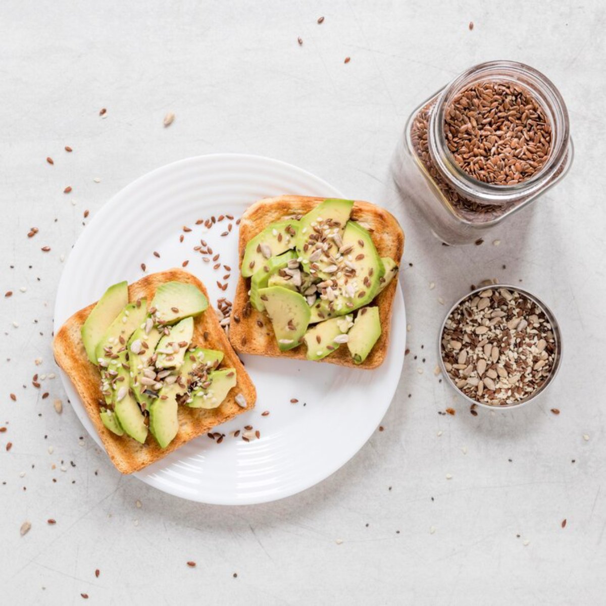 Avocado slices on toast with seeds, on a white plate, beside jars of flaxseeds on a light background.