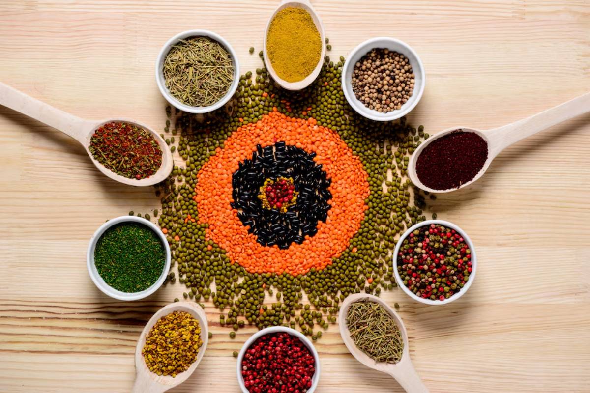 Various spices and lentils arranged in a circular pattern on a wooden surface, surrounded by spoons filled with colorful spices.