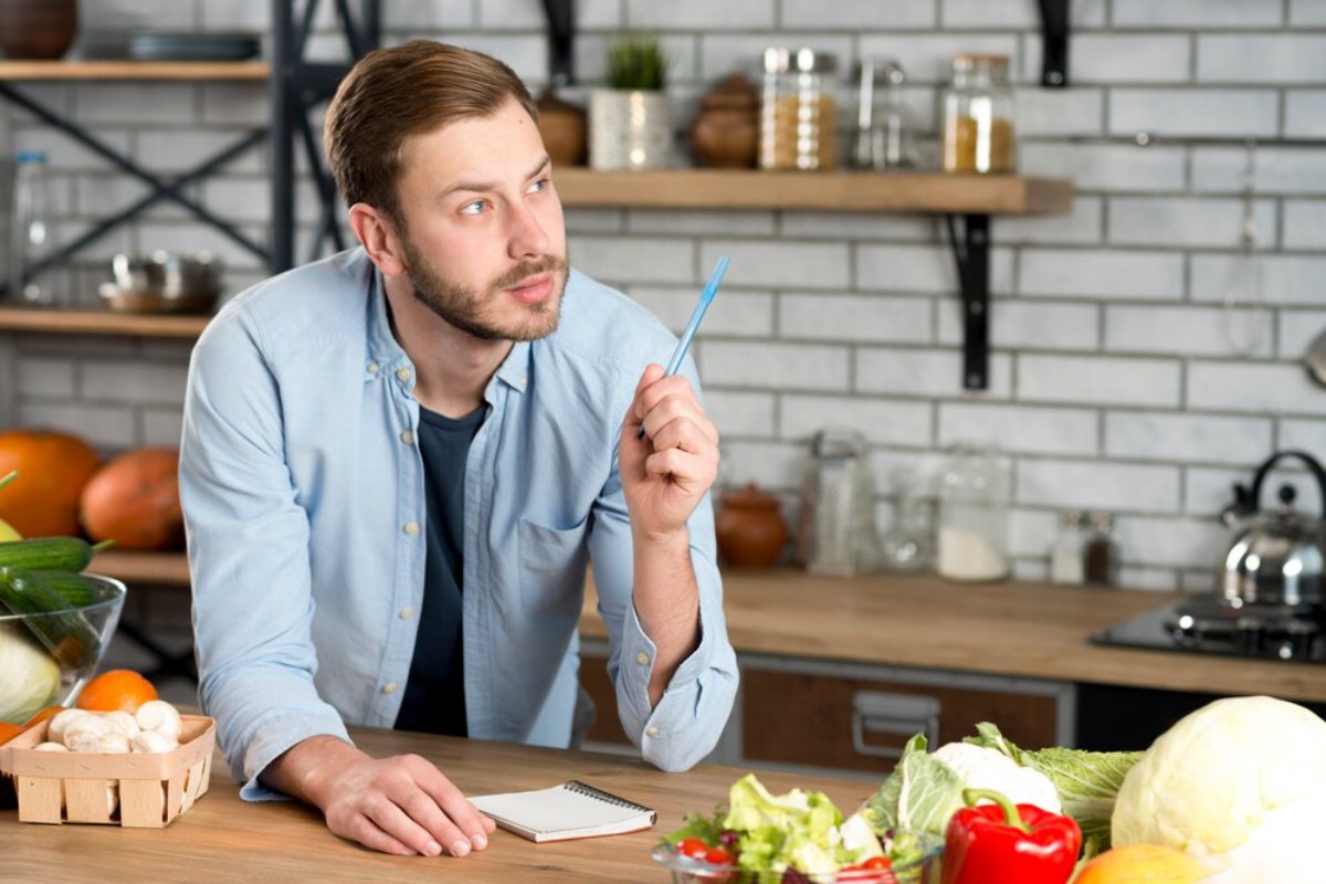 Man in a kitchen holding a pen and looking thoughtful, surrounded by fresh vegetables.