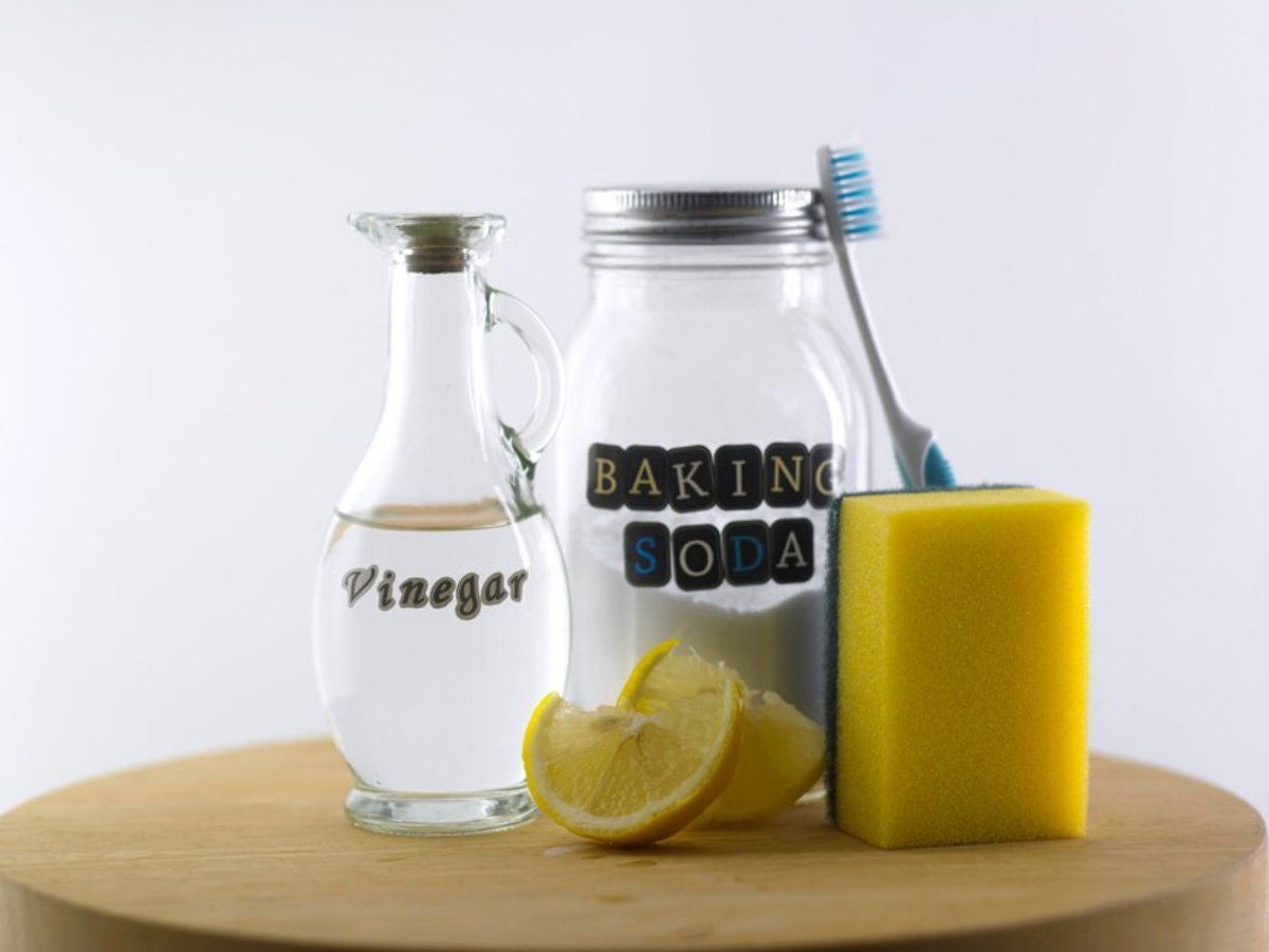 A jug of vinegar, jar of baking soda, toothbrush, sponge, and lemon slices on a wooden table.