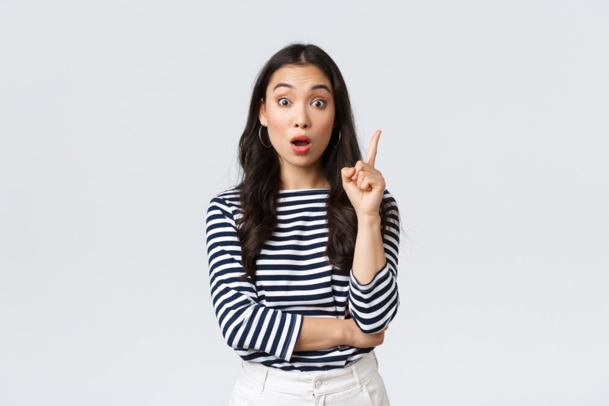 Surprised young woman in a striped shirt pointing upwards, isolated on a light background.