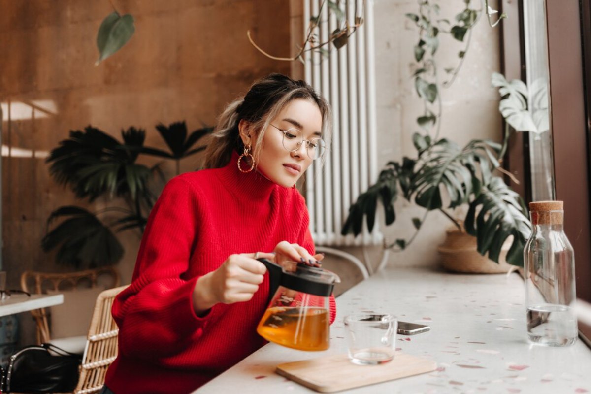 Young woman in glasses and red sweater pouring tea in a cafe with green plants in the background.