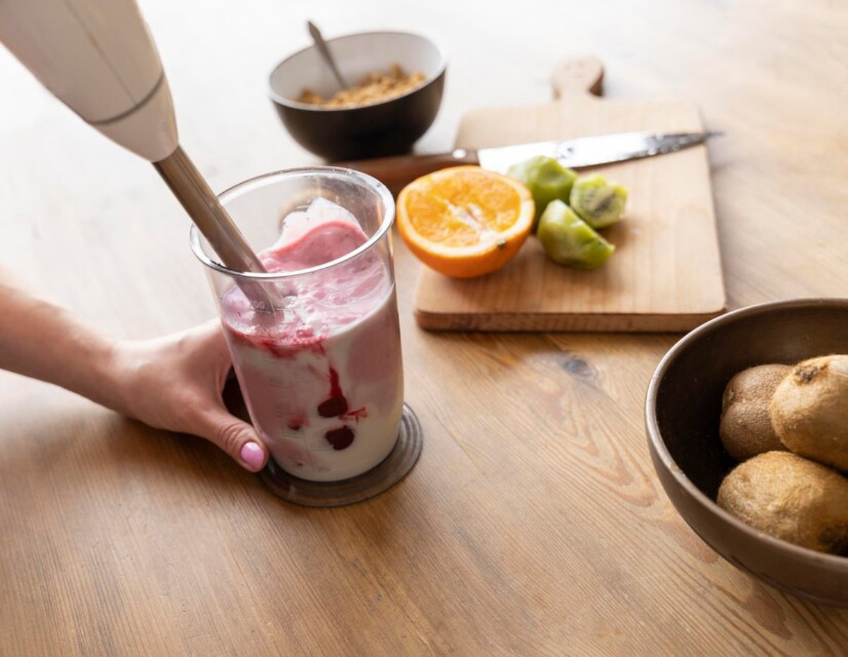 A hand using an immersion blender to mix a fruit smoothie in a glass cup on a wooden table.