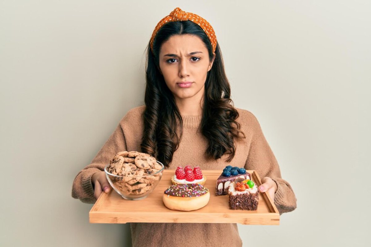 Woman holding a tray with cookies and assorted desserts, looking confused or undecided.