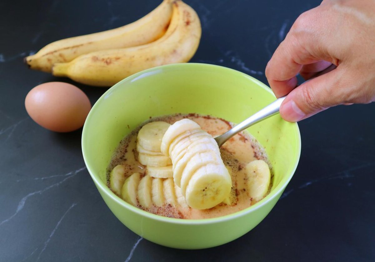 A bowl of oatmeal with sliced bananas being stirred, accompanied by eggs and whole bananas on a dark countertop.