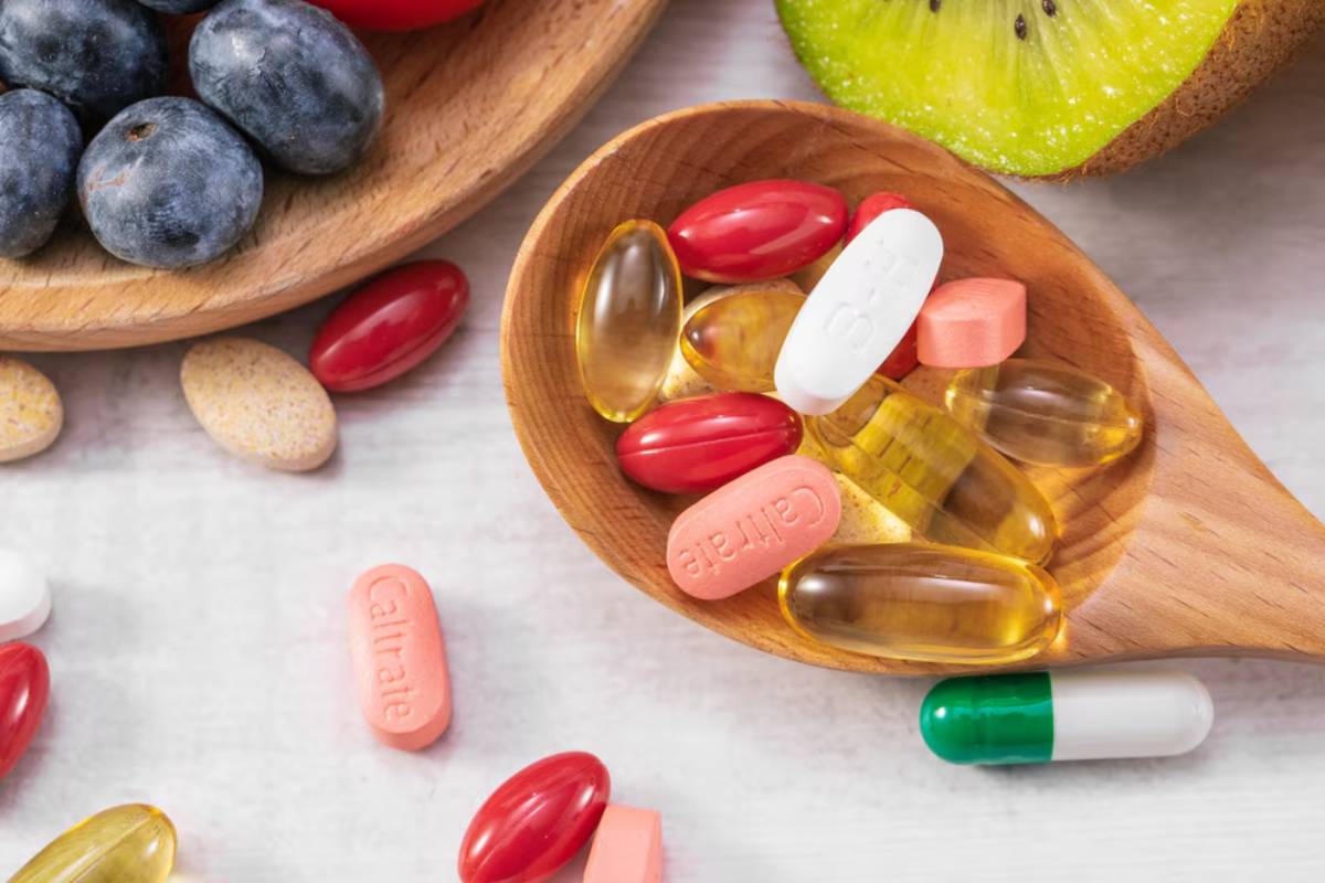 Assorted dietary supplements in a wooden spoon, next to fresh fruits on a marble surface.