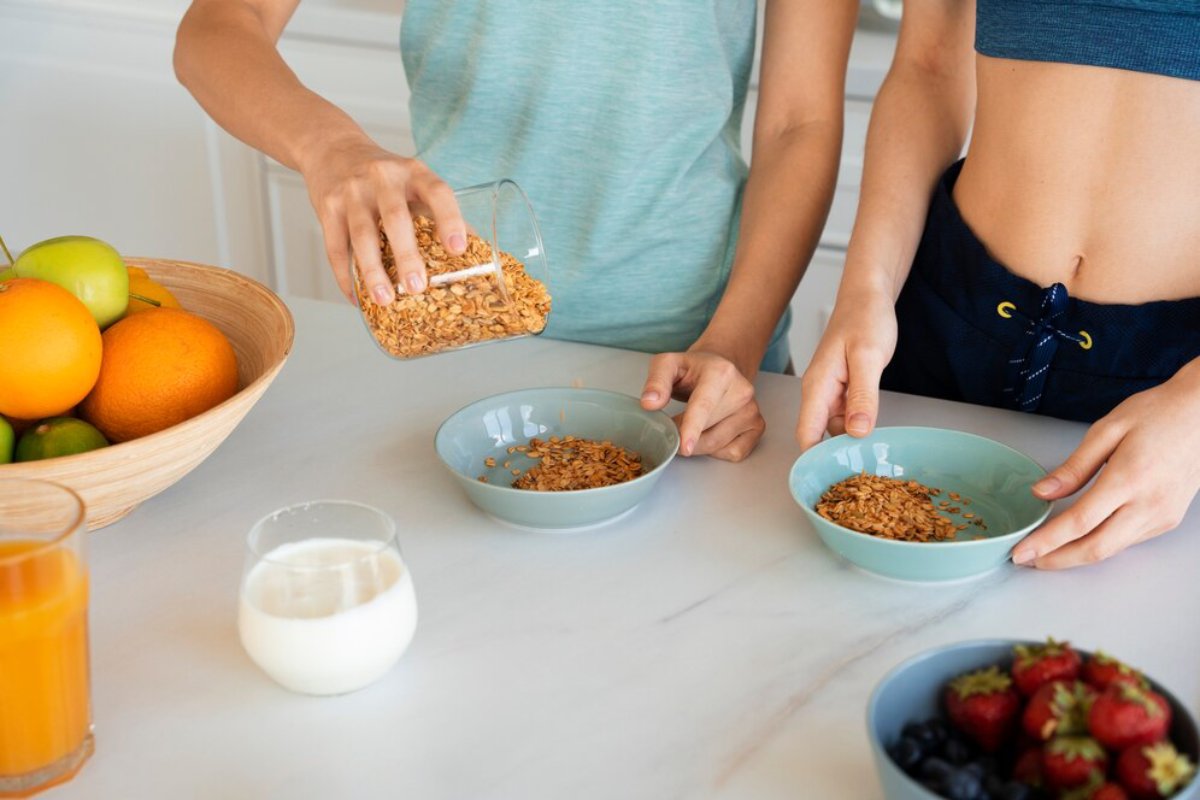 Two people pouring granola into bowls on a kitchen counter with fresh fruits and drinks nearby.
