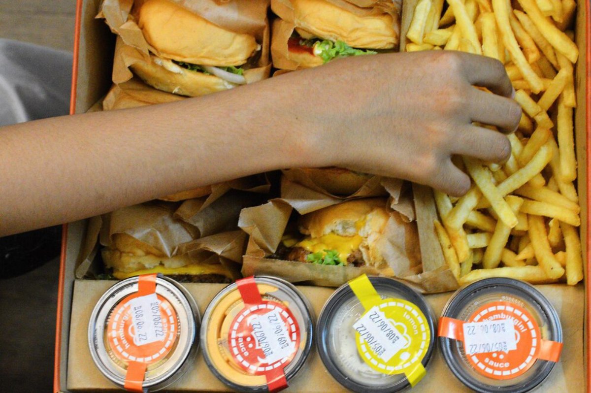 Hands reaching for burgers and fries in a takeaway box with condiment cups.