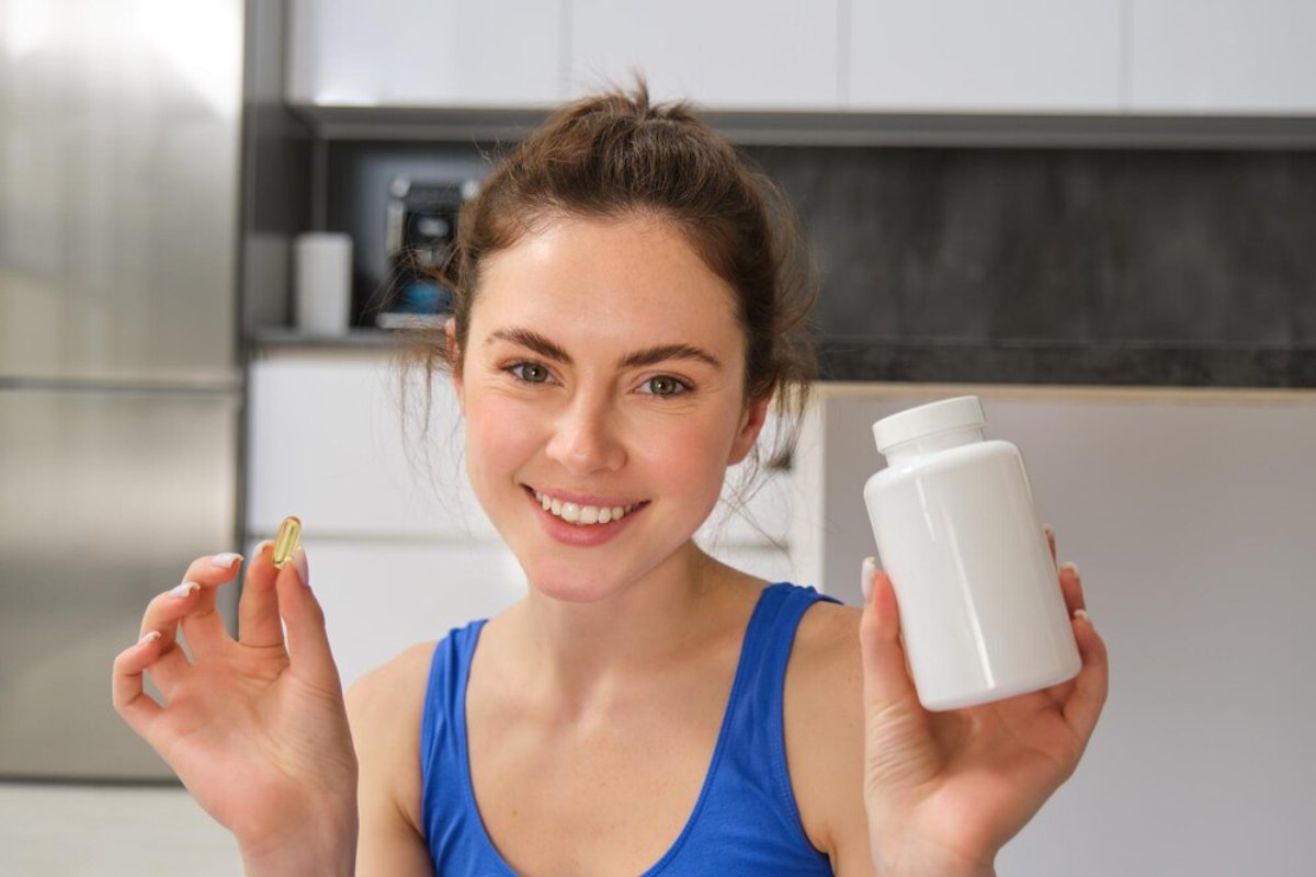 A smiling young woman holding a nutritional supplement capsule and a bottle in a modern kitchen.