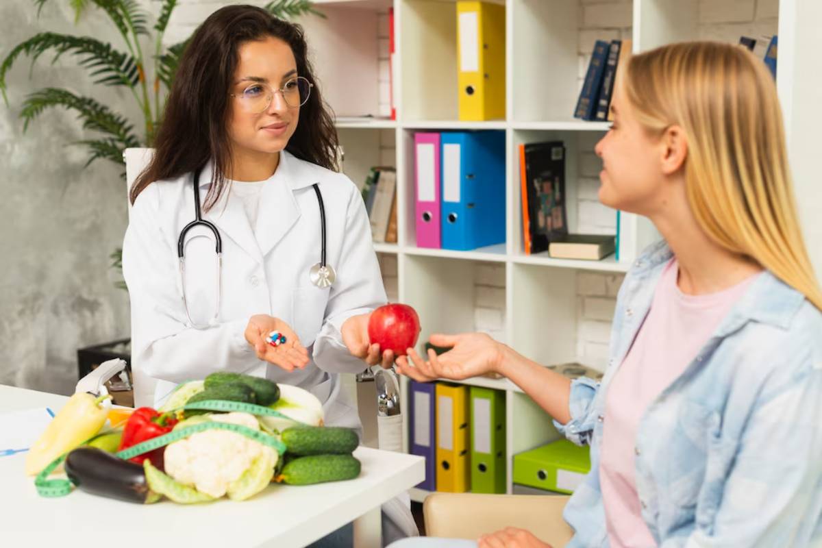 A female doctor hands a red apple to a female patient, with vegetables on the table in a clinic.