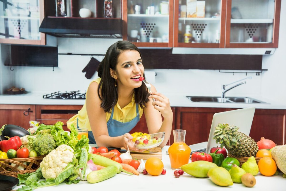 Woman eating fruit in a kitchen filled with fresh vegetables and fruits, with a laptop nearby.