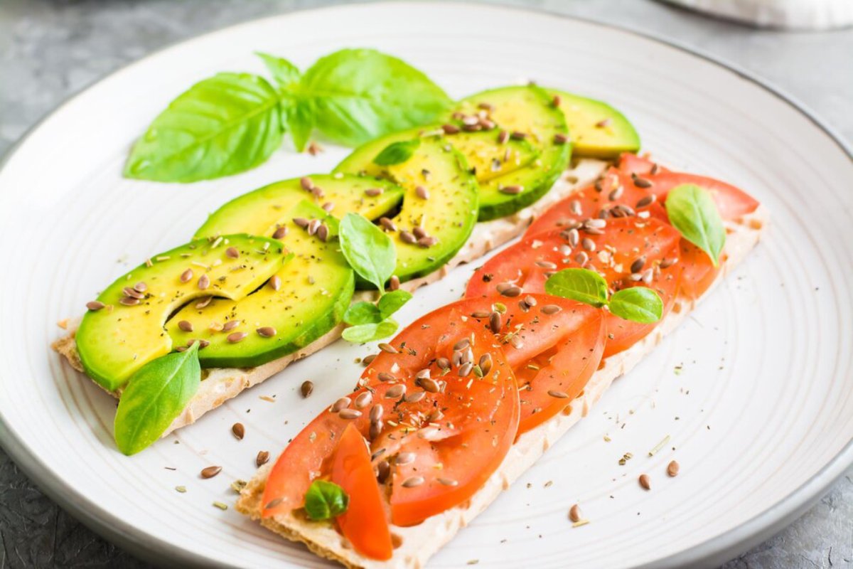 Avocado and tomato slices on bread, garnished with basil leaves and seeds on a white plate.