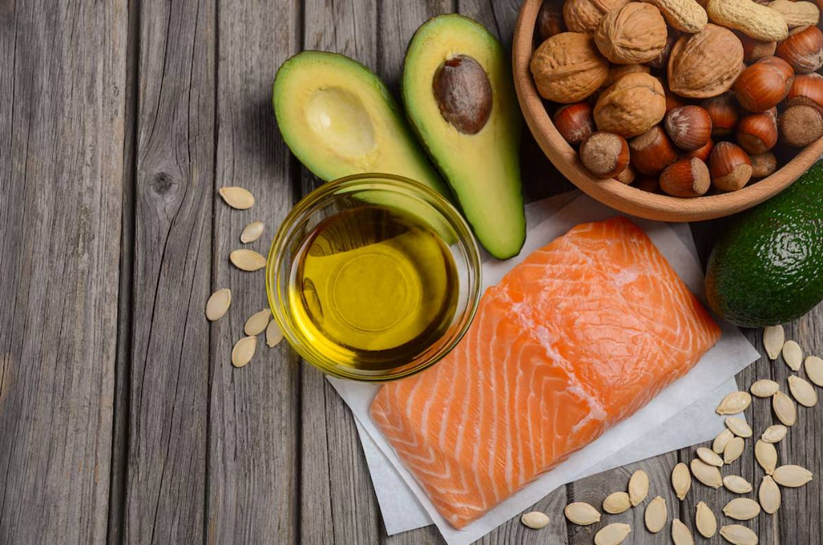 A variety of healthy foods including salmon, avocados, olive oil, and mixed nuts displayed on a wooden surface.