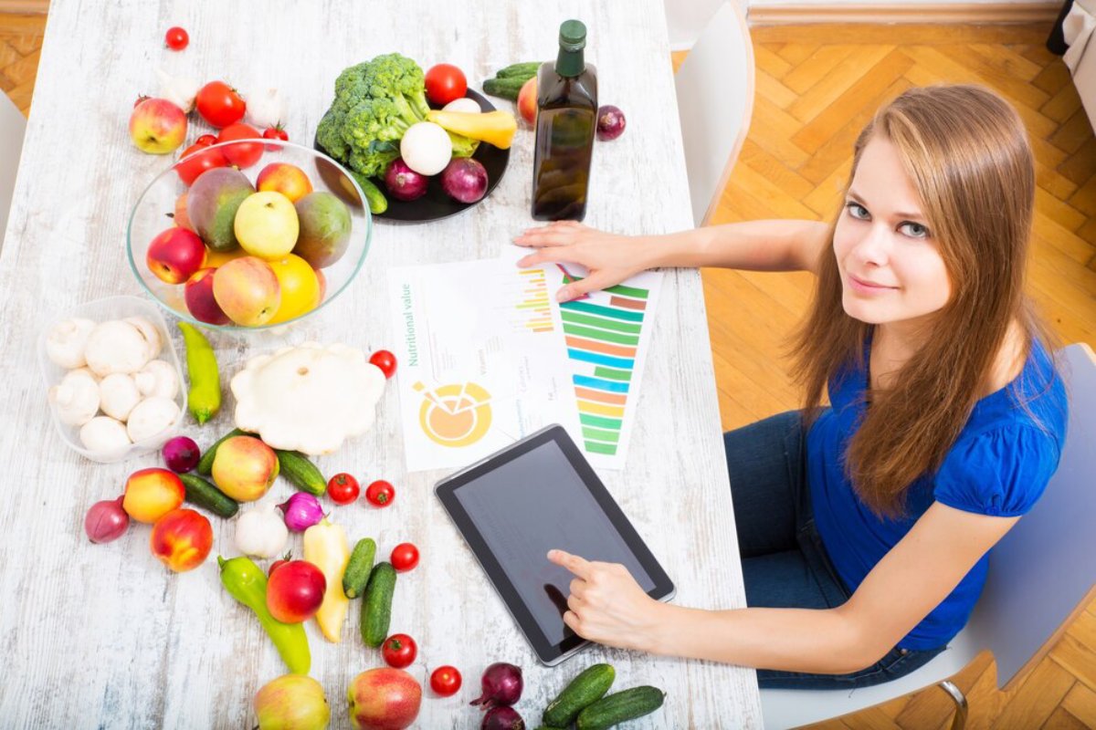 Woman sitting at table with fresh vegetables and fruits, analyzing nutritional charts on digital tablet.