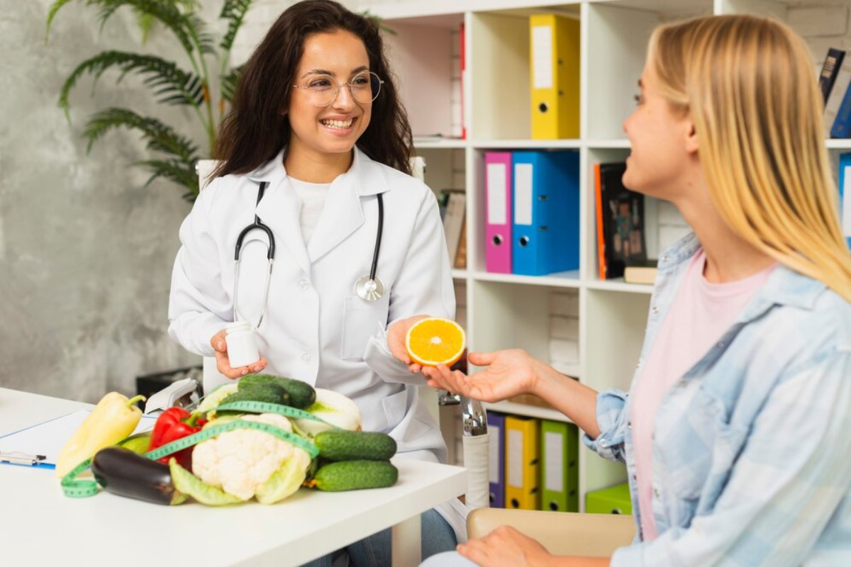 Female doctor discussing nutrition with a patient, holding a half orange, surrounded by vegetables in a clinic.
