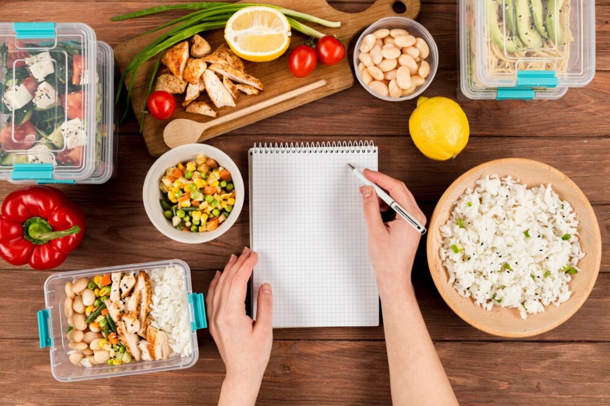 A hands-on meal prep scene with containers of salad, chicken, vegetables, and a notepad for planning meals. Fresh ingredients surround.