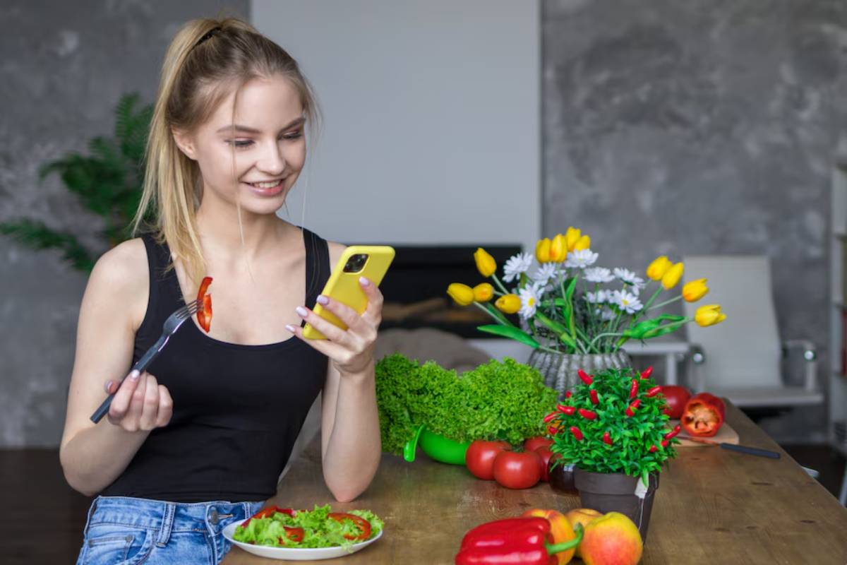 Young woman smiling at her phone while holding a fork with a slice of pepper, surrounded by fresh vegetables on a kitchen table.