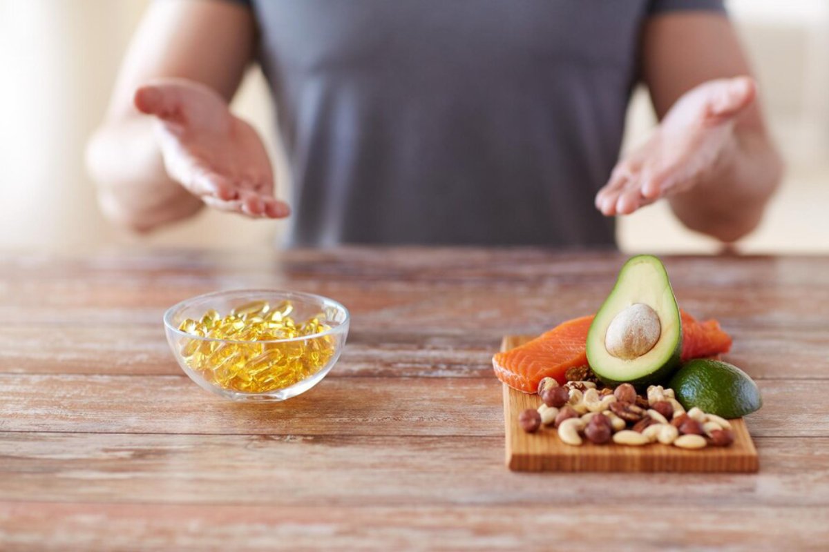 Person gesturing towards healthy foods including avocado, salmon, nuts, and omega-3 capsules on a wooden table.