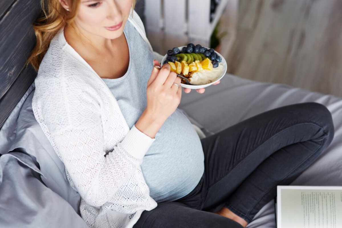 Pregnant woman sitting on couch, eating fruit-topped yogurt from a bowl.