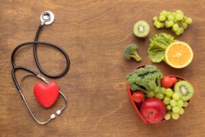 A stethoscope with a red heart rests on a wood surface next to a heart-shaped bowl filled with fresh vegetables and fruits.