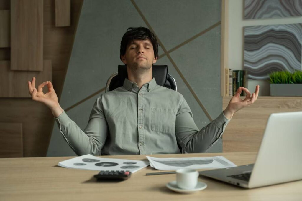 Man meditating at his desk in an office, surrounded by a laptop, papers, and a coffee cup.