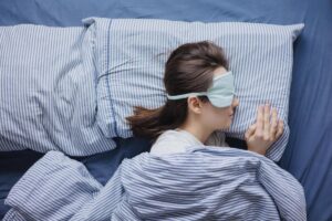 Woman sleeping in bed with a sleep mask, wearing striped pajamas and lying on blue sheets.