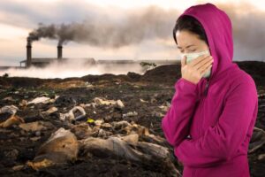 Woman in pink hoodie covers her face with a mask against industrial pollution and waste background.